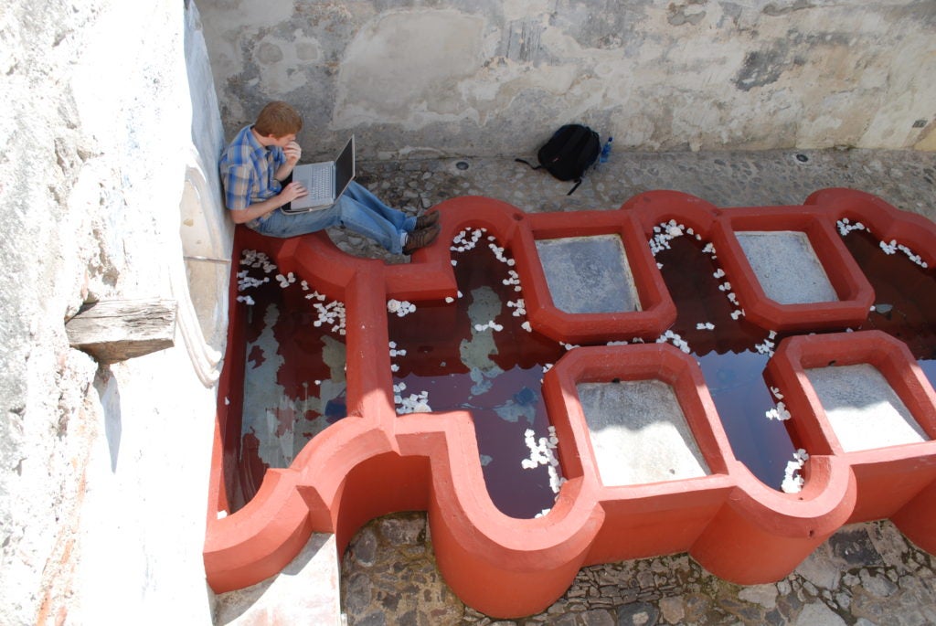 student looking at laptop while sitting on unique red painted fountain pool architecture in stone courtyard