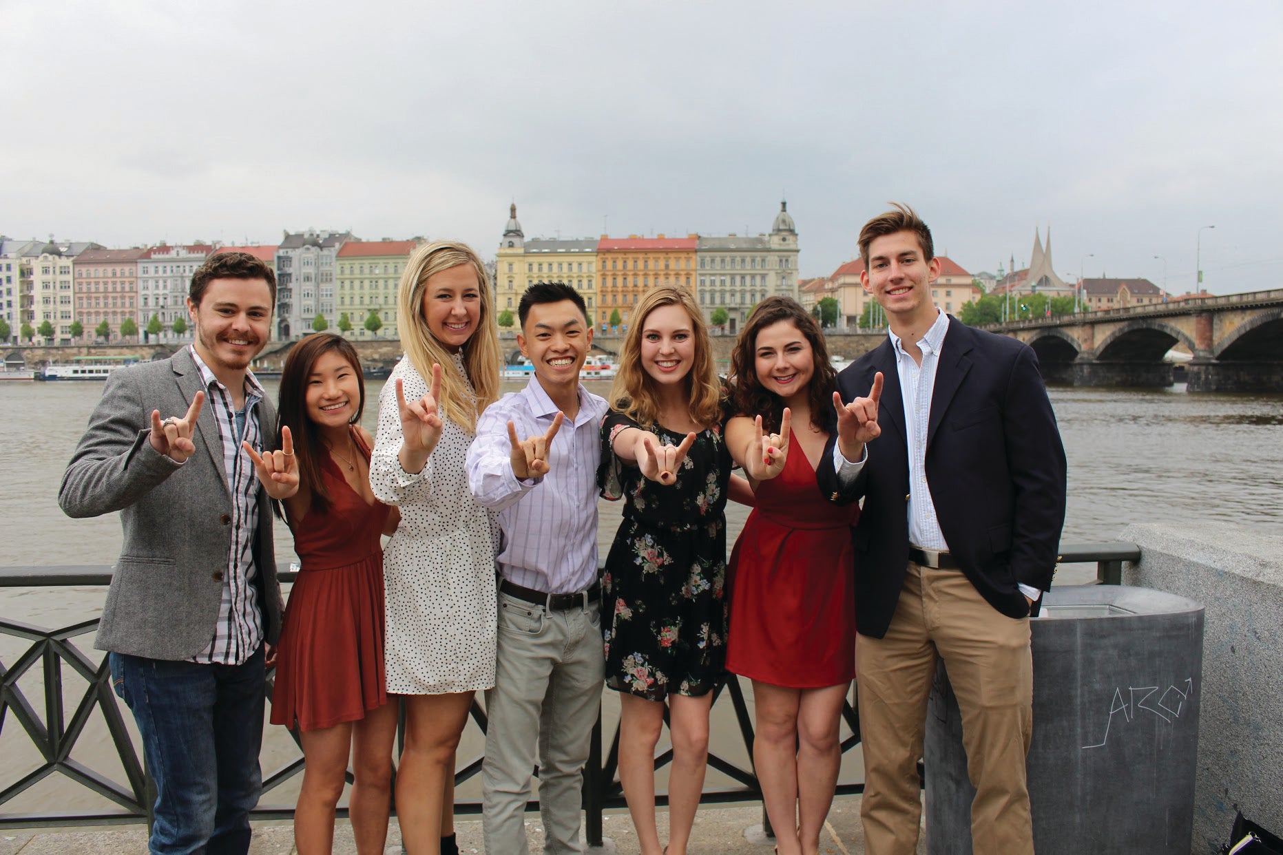 A diverse mix of UT abroad students pose with the Hook 'Em horns on a pier on a rivers edge with buildings in the background